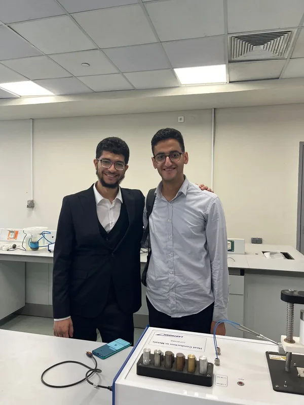 Ahmed in a suit with a colleague in a university lab, standing behind a heat-conduction experiment