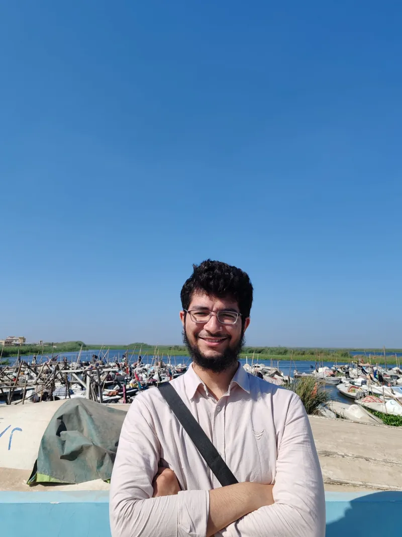 Ahmed standing with arms crossed by a harbour full of small fishing boats under a clear blue sky